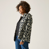 A woman with curly dark hair is shown in profile wearing a black and cream floral patterned jacket over a black top and blue jeans, photographed against a plain light gray studio background.