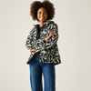 A woman with an afro hairstyle stands against a light gray studio background wearing a black and off-white floral patterned jacket, a black top, and blue jeans.
