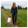 Woman walking on a path in a field holding a white bucket, wearing a black outfit with a visible brand logo.