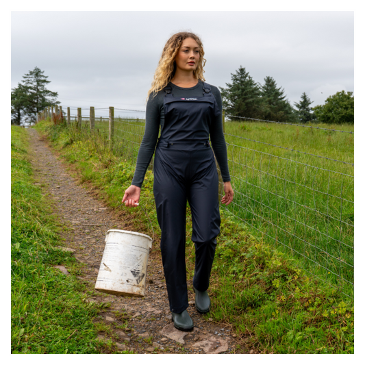 Woman walking on a path in a field holding a white bucket, wearing a black outfit with a visible brand logo.