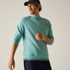 A man wearing a light teal-green crewneck sweatshirt with a small red and yellow logo on the left chest, paired with dark navy pants and a navy baseball cap, photographed against a neutral light beige background.