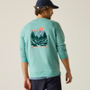 A man wearing a mint green crewneck sweatshirt with a large back graphic of a mountain landscape and pine trees, paired with a dark blue cap and navy pants, photographed against a solid light beige background.
