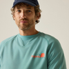 A close-up portrait of a man wearing a teal crewneck sweatshirt with a small orange and white logo on the left chest, paired with a navy blue cap, against a plain light beige background.