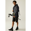 A man modeling a dark gray waterproof hooded jacket, black hiking shorts, and black hiking boots while holding a trekking pole against a light gray studio background.