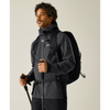 A man wearing a dark gray outdoor jacket and a black backpack, holding a trekking pole against a plain light gray studio background.