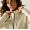 A close-up of a woman wearing a cream-colored zip-up hoodie, pulling the zipper with both hands. The background is a clean, neutral off-white studio setting.