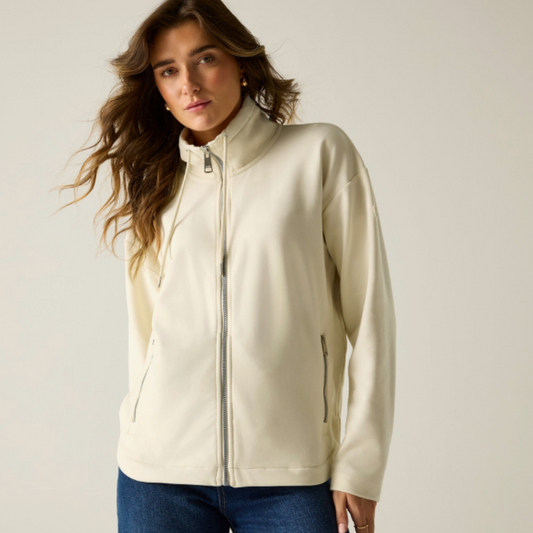 A woman wearing a cream-colored zip-up jacket with a mock neck and side pockets, paired with blue jeans, photographed against a neutral light beige background.