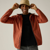 A smiling man wearing a rust-orange lightweight zip-front jacket over a black shirt and gray jeans, with a beige bucket hat, posing against a light gray studio background.