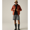 A man modeling an orange-brown hooded jacket over a black t-shirt, paired with gray denim shorts, black rain boots, and a beige bucket hat, against a solid light gray studio background.