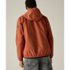 A person shown from the back wearing a hooded jacket in a muted terracotta or burnt orange color, paired with gray jeans, against a plain light gray studio background.
