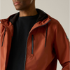 Close-up of a man wearing a rust-orange hooded jacket over a black shirt, with the jacket's front zipper, chest zip pocket, and drawstrings visible against a plain light beige background.