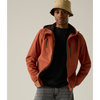 A man wearing a rust-orange hooded jacket over a black shirt, paired with gray jeans and a beige bucket hat, standing against a solid light gray background.