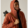A man wearing a hooded rust-orange waterproof jacket over a black shirt, holding a hiking pole and wearing a backpack, photographed against a plain light beige studio background.