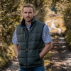A man standing on a dirt path in a wooded outdoor setting, wearing a dark green quilted puffer vest over a blue and white checkered short-sleeve shirt and blue jeans.