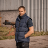 A man wearing a navy blue sleeveless puffer vest over a dark t-shirt, standing outdoors in front of a corrugated metal building.