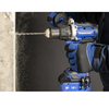 A close-up action shot of a person wearing blue and black work gloves and a dark jacket, using a blue and gray cordless drill to drill into a concrete wall, with dust particles flying.