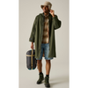 A man wearing an olive green hooded raincoat, a tan bucket hat, a brown plaid shirt, denim shorts, and hiking boots, holding a dark gray and tan packed sleeping bag against a plain beige studio background.