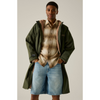 A male model wearing an olive green hooded jacket with a tan fleece lining, layered over a brown and beige plaid button-down shirt and light-wash denim shorts, set against a plain beige studio background.