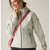 A woman wearing a light coloured hooded softshell jacket with a pink crossbody bag, posing against a solid light beige background.