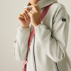 A close-up of a person wearing a light gray hooded jacket with a pink shoulder strap and a small black logo patch on the sleeve, against a plain light gray background.