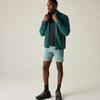 A man modeling a teal green lightweight zip-up jacket over a black t-shirt, paired with light blue running shorts, black socks, and black trail running shoes, against a solid light beige studio background.
