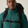 A man wearing a teal fleece zip-up jacket with a black backpack and harness straps over his shoulders, photographed against a plain light beige studio background.