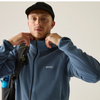 A man wearing a slate blue fleece zip-up jacket and a black cap, with a black shoulder bag visible on his back, set against a plain light background.