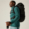 A man wearing a teal fleece pullover and light blue pants carries a large black hiking backpack on his back, holding trekking poles against a plain light gray studio background.