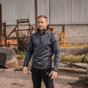 A man wearing a dark gray heathered quarter-zip pullover and black cargo pants stands in an industrial outdoor setting with scrap metal and a corrugated metal wall in the background.