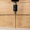 A close-up of a gold-colored wood screw being driven into a light wood plank by a drill, set against a background of stacked wooden planks.