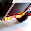 Close-up of a black trailer or utility vehicle rear corner with two illuminated amber reflector lights and a visible tire on a light gray floor.