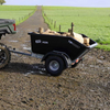 A black TipAssist Agri trailer loaded with logs, attached to a vehicle on a muddy country road with green fields on both sides.