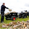 A man loads wood logs into a black agricultural trailer hitched to a black quad bike on a grassy field. The trailer has white branding that reads 'SHOT AGRI'.
