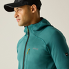 A man in side profile wearing a teal hooded athletic jacket with a black zipper and a black baseball cap, photographed against a plain light beige studio background.