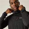 A man wearing a black zip-up jacket with red zipper pulls and a white logo on the chest, posing with his hands on the collar against a light beige background.