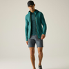 A male model wearing a teal green lightweight zip-up hooded jacket over a matching teal long-sleeve shirt, gray running shorts, and a black baseball cap, standing against a plain light gray studio background.