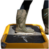 A person wearing dark green rubber boots steps into a yellow and black cleaning pad or mat, with a second boot standing beside it on a white background.