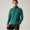 A man wearing a teal green quarter-zip fleece pullover with a small black logo on the left chest, paired with dark gray pants, standing against a light gray studio background.