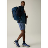 A man wearing a blue technical backpack, a navy quilted vest over a dark long-sleeve shirt, light blue shorts, and blue athletic socks and running shoes, 