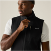 A man wearing a black zip-front vest over a white short-sleeve shirt, photographed against a plain light beige studio background. The vest has a stand collar, a front zip closure, a vertical zip chest pocket