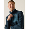 A smiling man wearing a dark blue quarter-zip fleece pullover with a teal chest pocket and a visible Patagonia logo patch, photographed against a plain light beige studio background.