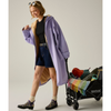 A woman models a lavender hooded coat with a tan fur-trim over a black top and denim shorts, standing beside a black REGATTA shopping cart loaded with colorful items, against a plain light gray studio background.