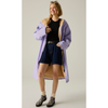 A model is wearing a lavender hooded coat with a tan fleece lining, styled over a black top, dark denim shorts, cream socks, and black shoes with a purple pattern, standing against a neutral studio background.