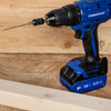 A blue and black PRAWLPLUG cordless drill with a wood screw bit, resting on a wooden surface with a wood plank in the foreground.