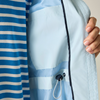 Close-up of a hooded jacket in a light warm gray or off-white color, featuring a pink zipper trim along the collar and a small black rectangular logo patch on the upper sleeve.