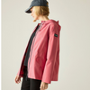 A woman wearing a pink hooded rain jacket with a black zip closure, layered over a black top and pants, and wearing a dark baseball cap, photographed against a plain light gray studio background.