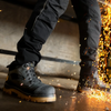 A close-up of a person wearing black work boots and black pants, standing on a concrete floor with sparks flying near the right side.