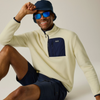 A man wearing a cream quarter-zip fleece pullover with a navy chest pocket, navy shorts, a navy bucket hat, and blue mirrored sunglasses, posing against a light beige studio background.