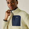 A close-up of a man wearing a cream-colored fleece pullover with a navy blue chest pocket and zipper detail. The brand name is visible on a small black label on the pocket.