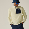 A man wearing a cream-colored quarter-zip fleece pullover with a navy chest pocket, paired with navy pants, a navy bucket hat, and blue sunglasses, set against a plain light beige background.